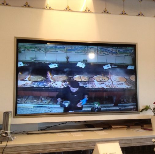 A TV showing 7 paella pans cooking atop wood burning stoves, and the cook in the process of making another.